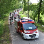 Na pierwszym planie czerwony samochód strażacki za nim peleton rowerów. Po obu stronach ulicy zielone drzewa i trawniki