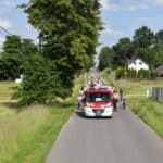 Z górki zjeżdża peleton rowerzystów, prowadzi ich wóz strażacki. Z boku rośnie wysokie drzewo, w tle stoi biały dom