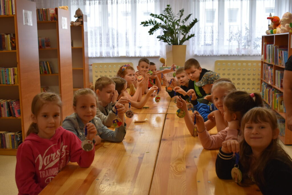 Uśmiechnięte dzieci siedzą przy stole i trzymają breloczki, za nimi regały z książkami