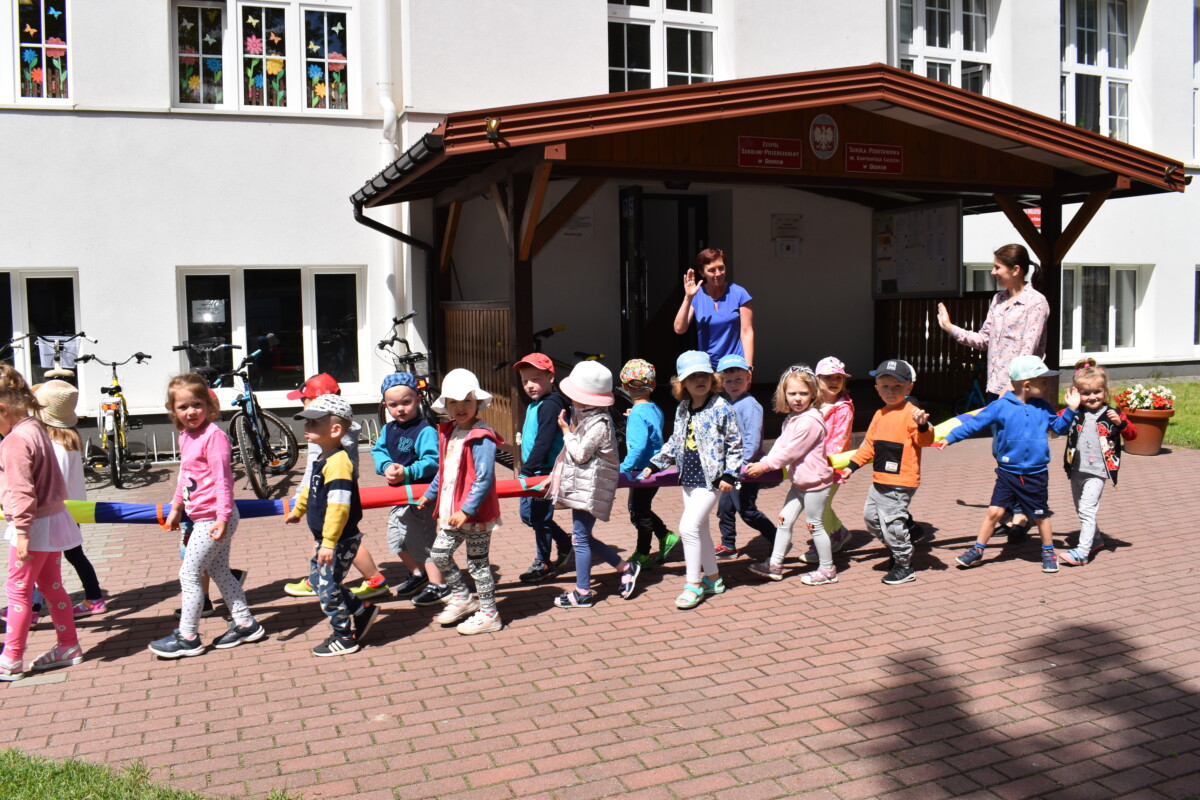 Grupka dzieci idzie w parach machają ręką dwie panie. W tle biały budynek i drewniany ganek.