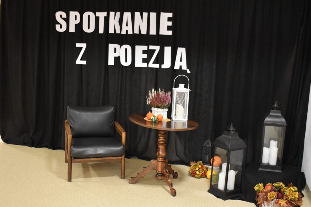 Czarna kotara na niej biały napis Spotkanie z poezją, przed kotarą leżą lampiony, dynie, fotel, stolik na którym stoi wrzos, tykwy i lampion