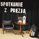 Czarna kotara na niej biały napis Spotkanie z poezją, przed kotarą leżą lampiony, dynie, fotel, stolik na którym stoi wrzos, tykwy i lampion
