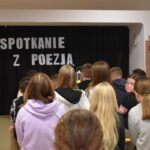 Dzieci siedzą na ławkach, przed nimi stoi kobieta. W tle czarna kotara na niej napis Spotkanie z poezją