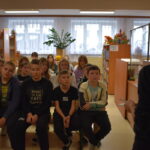 Na ławkach siedzi grupa dzieci i słucha kobiety, która stoi przed nimi. W tle regały z książkami