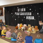 Grupa przedszkolaków i dwie panie pozują do zdjęcia. Dzieci w rękach trzymają kolorowe misie. W tle czarna kotara z białym napisem i gwiazdkami, przed nimi leżą kolorowe poduszki