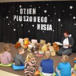 Grupa przedszkolaków i dwie panie pozują do zdjęcia. Dzieci w rękach trzymają kolorowe misie. W tle czarna kotara z białym napisem i gwiazdkami, przed nimi leżą kolorowe poduszki