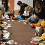 Grupa przedszkolaków i dwie panie pozują do zdjęcia. Dzieci w rękach trzymają kolorowe misie. W tle czarna kotara z białym napisem i gwiazdkami, przed nimi leżą kolorowe poduszki