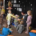 Przedszkolaki siedzą na kolorowych poduszkach i oglądają książki przed nimi leży kilkanaście misiów pluszowych w tle czarna kotara z białym napisem