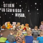 Przedszkolaki siedzą na kolorowych poduszkach i oglądają książki przed nimi leży kilkanaście misiów pluszowych w tle czarna kotara z białym napisem