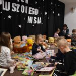 Przedszkolaki siedzą na kolorowych poduszkach i oglądają książki przed nimi leży kilkanaście misiów pluszowych w tle czarna kotara z białym napisem
