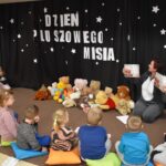 Przedszkolaki siedzą na kolorowych poduszkach i oglądają książki przed nimi leży kilkanaście misiów pluszowych w tle czarna kotara z białym napisem