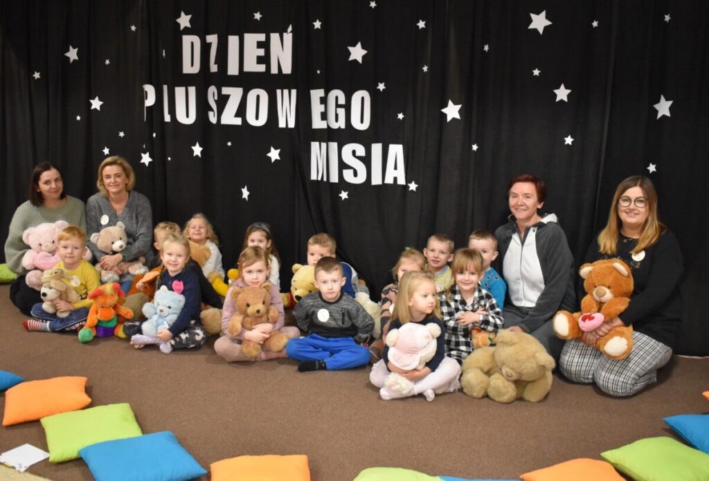 Przedszkolaki siedzą na kolorowych poduszkach i oglądają książki przed nimi leży kilkanaście misiów pluszowych w tle czarna kotara z białym napisem