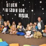 Przedszkolaki siedzą na kolorowych poduszkach i oglądają książki przed nimi leży kilkanaście misiów pluszowych w tle czarna kotara z białym napisem