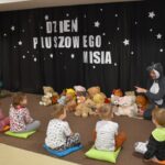 Przedszkolaki siedzą na kolorowych poduszkach i oglądają książki przed nimi leży kilkanaście misiów pluszowych w tle czarna kotara z białym napisem