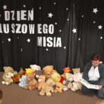 Przedszkolaki siedzą na kolorowych poduszkach i oglądają książki przed nimi leży kilkanaście misiów pluszowych w tle czarna kotara z białym napisem