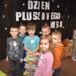 Przedszkolaki siedzą na kolorowych poduszkach i oglądają książki przed nimi leży kilkanaście misiów pluszowych w tle czarna kotara z białym napisem