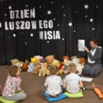 Przedszkolaki siedzą na kolorowych poduszkach i oglądają książki przed nimi leży kilkanaście misiów pluszowych w tle czarna kotara z białym napisem
