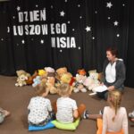 Przedszkolaki siedzą na kolorowych poduszkach i oglądają książki przed nimi leży kilkanaście misiów pluszowych w tle czarna kotara z białym napisem