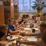 Przy dużym drewnianym stole siedzą dzieci i dekorują pierniczki kolorowymi posypkami, po bu stronach stoją regały z kolorowymi książkami