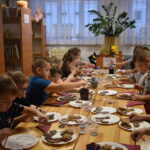 Przy dużym drewnianym stole siedzą dzieci i dekorują pierniczki kolorowymi posypkami, po bu stronach stoją regały z kolorowymi książkami