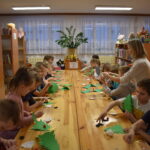 Przy dużym drewnianym stole siedzą dzieci i dekorują pierniczki kolorowymi posypkami, po bu stronach stoją regały z kolorowymi książkami