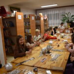 Przy dużym drewnianym stole siedzą dzieci i dekorują pierniczki kolorowymi posypkami, po bu stronach stoją regały z kolorowymi książkami