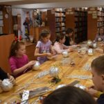 Przy dużym drewnianym stole siedzą dzieci i dekorują pierniczki kolorowymi posypkami, po bu stronach stoją regały z kolorowymi książkami