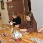 Przy dużym drewnianym stole siedzą dzieci i dekorują pierniczki kolorowymi posypkami, po bu stronach stoją regały z kolorowymi książkami