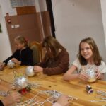 Przy dużym drewnianym stole siedzą dzieci i dekorują pierniczki kolorowymi posypkami, po bu stronach stoją regały z kolorowymi książkami