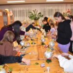 Przy dużym drewnianym stole siedzą dzieci i dekorują pierniczki kolorowymi posypkami, po bu stronach stoją regały z kolorowymi książkami