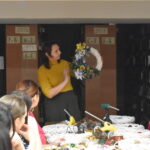 348 Kobieta trzyma w ręku ozdobę świąteczną na drzwi, resztę pań przygląda się jej. W tle regały z książkami.