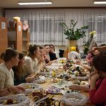 367 W pomieszczeniach Biblioteki Publicznej odbywają się warsztaty robienia wianków na drzwi. Instruktorka pokazuje zrobiony już wianek. W tle okna duży kwiatek oraz regały z książkami.