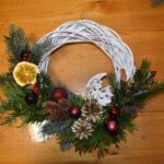 437 Pleciony biały wianek ozdobiony czarnymi bombkami, zielonymi gałązkami świerku, tui, szyszkami, cynamonem i pomarańczą.