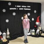 030 Dziewczynka w różowym sweterku śpiewa do mikrofonu. W tle czarny materiał na którym widnieje napis II Gminny Przegląd Kolęd, Pastorałek i Piosenek świątecznych. Na podłodze stoją kolorowe skrzaty, lampiony i pieńki.