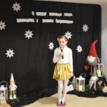 030 Dziewczynka w różowym sweterku śpiewa do mikrofonu. W tle czarny materiał na którym widnieje napis II Gminny Przegląd Kolęd, Pastorałek i Piosenek świątecznych. Na podłodze stoją kolorowe skrzaty, lampiony i pieńki.