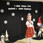 036 W białej bluzce i złotej spódnicy stoi dziewczynka w reku ma mikrofon. W tle czarny materiał na którym widnieje napis II Gminny Przegląd Kolęd, Pastorałek i Piosenek świątecznych. Na podłodze stoją kolorowe skrzaty, lampiony i pieńki.