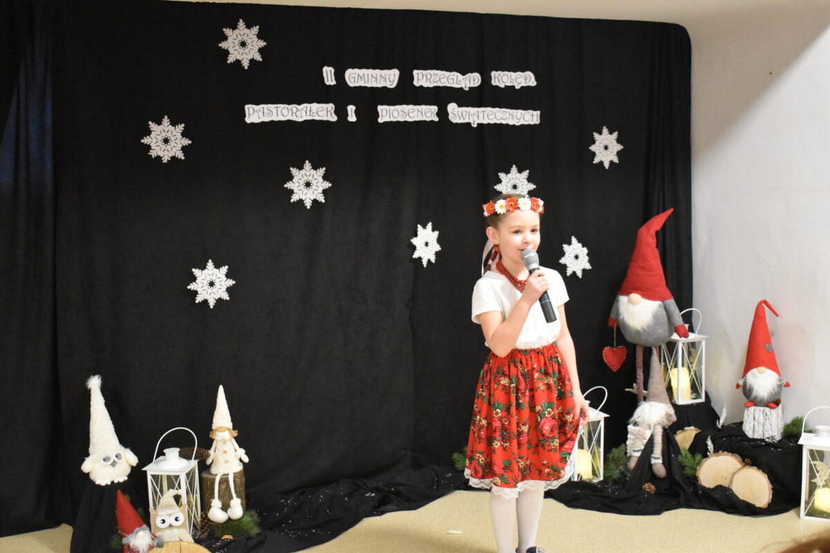 036 W białej bluzce i złotej spódnicy stoi dziewczynka w reku ma mikrofon. W tle czarny materiał na którym widnieje napis II Gminny Przegląd Kolęd, Pastorałek i Piosenek świątecznych. Na podłodze stoją kolorowe skrzaty, lampiony i pieńki.