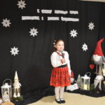 064 Chłopiec ubrany na niebiesko w białej koszuli trzyma mikrofon w ręku. W tle czarny materiał na którym widnieje napis II Gminny Przegląd Kolęd, Pastorałek i Piosenek świątecznych. Na podłodze stoją kolorowe skrzaty, lampiony i pieńki.