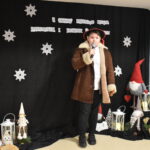 083 Młoda dziewczyna w czarnej sukience trzyma mikrofon w ręku, na tle czarnej kotary na której są białe gwiazdki i napis, oraz kolorowe skrzaty.