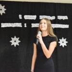 150 Młoda kobieta w białej bluzce trzyma mikrofon. W tle czarna kotara z białymi gwiazdkami i napisem.