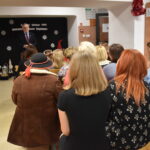 083 Młoda dziewczyna w czarnej sukience trzyma mikrofon w ręku, na tle czarnej kotary na której są białe gwiazdki i napis, oraz kolorowe skrzaty.