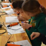 W bibliotece na zielonym dywanie siedzą dzieci w kółku. W tle regały z książkami.