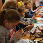 Przy dużym stole dzieci robią kanapki, smarują masłem chleb, kroją wędlinę. Na stole stoi srebrna okrągła taca.