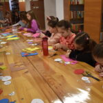 145 W bibliotece przy długim drewnianym stole na którym leżą nożyczki, ołówki i ścinki kolorowych kartek siedzą dzieci. Jedna dziewczynka podnosi rękę, jedna wycina. W tle dwa okna i regały z kolorowymi książkami.