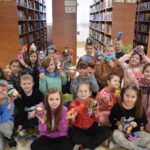 160 Uśmiechnięte dzieci pozują do zdjęcia z zrobionymi przez siebie skrzatami i magnesami. W tle biblioteczne regały z książkami.