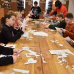 168 Dzieci zgromadzone przy dwóch stołach na których stoi miska wypełniona mąką i drożdżarzami oraz dzbanek z wodą.