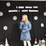 127 Panienka w czarnej sukience z białym kołnierzykiem trzyma mikrofon w ręku. w tle czarna kotara z białymi gwiazdkami i napisem za nią stoi skrzat.