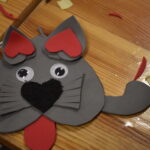 196 Grupka dzieci siedzi na zielonym dywanie i pomarańczowych matach. Przed nimi siedzi kobieta w czarnej bluzce. W tle regały biblioteczne z książkami.