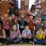 202 W pomieszczeniach biblioteki dzieci siedzą przy drewnianym stole i malują przezroczyste szklanki farbami trzymają pędzelki w dłoniach. W tle regały z książkami.