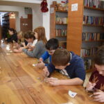 Dzieci siedzą przy stole na którym leżą kable i długopisy 3D w tle regały biblioteczne z książkami.